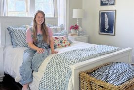 A smiling young woman in overalls sits on a neatly made bed with blue and white bedding in a bright bedroom with a wicker basket, floral pillow, and framed art on the wall.