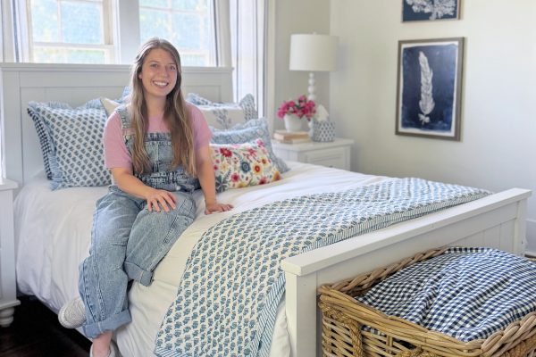 A smiling young woman in overalls sits on a neatly made bed with blue and white bedding in a bright bedroom with a wicker basket, floral pillow, and framed art on the wall.