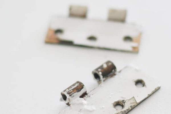 Two rusty, paint-chipped door hinge plates lying on a white surface, one in the foreground and one slightly out of focus in the background.