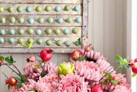 A white vase holds pink flowers, green artichokes, and red pomegranates. Behind it, an abacus with blue and green beads hangs on a white-paneled wall.