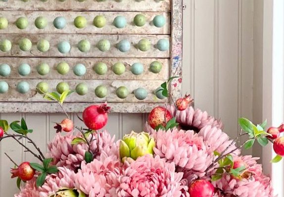 A white vase holds pink flowers, green artichokes, and red pomegranates. Behind it, an abacus with blue and green beads hangs on a white-paneled wall.