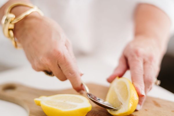 A person uses a spoon to scoop out a lemon half on a wooden cutting board, with another lemon half nearby. The person wears a white shirt and a gold bracelet.