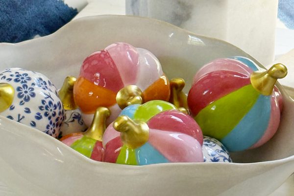 A white ceramic bowl holds small, glossy pumpkins with colorful pastel stripes and gold stems, alongside blue and white ceramic balls, resting on a white surface near a marble lamp base—creating a charming seasonal display that could inspire ideas for how to hang a wreath in your own home.