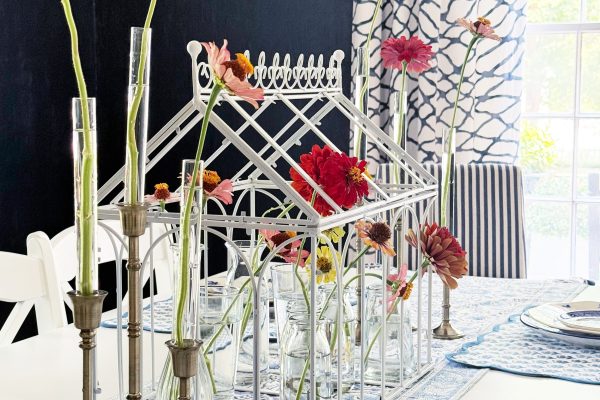 A white dining table with a metal greenhouse centerpiece holds red flowers in glass vases. Brass candlesticks with red blooms, blue patterned plates, and blue and white decor complete the scene, creating an inviting space that would perfectly complement your next home project—perhaps you’re considering How To Install Peel and Stick Wall Molding for a stylish finishing touch.