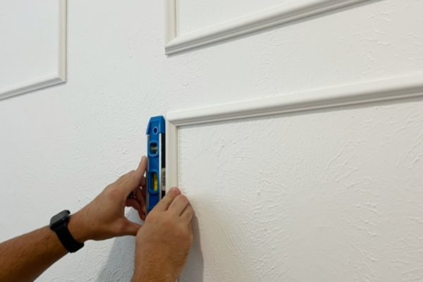 A person uses a blue spirit level to check the alignment of decorative trim on a textured white wall.