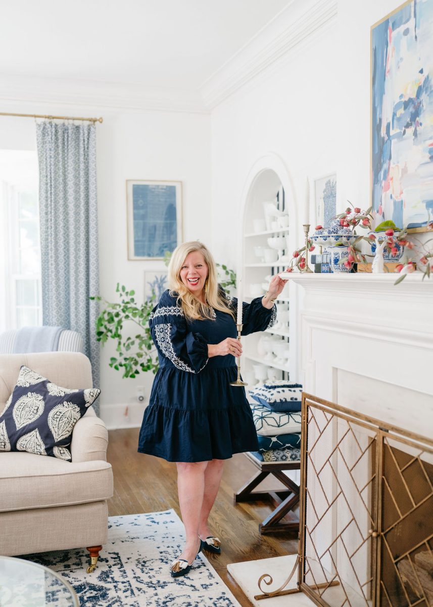 A woman in a navy dress stands smiling by a white fireplace, holding a champagne glass in a bright, stylish living room with blue and white decor accents.