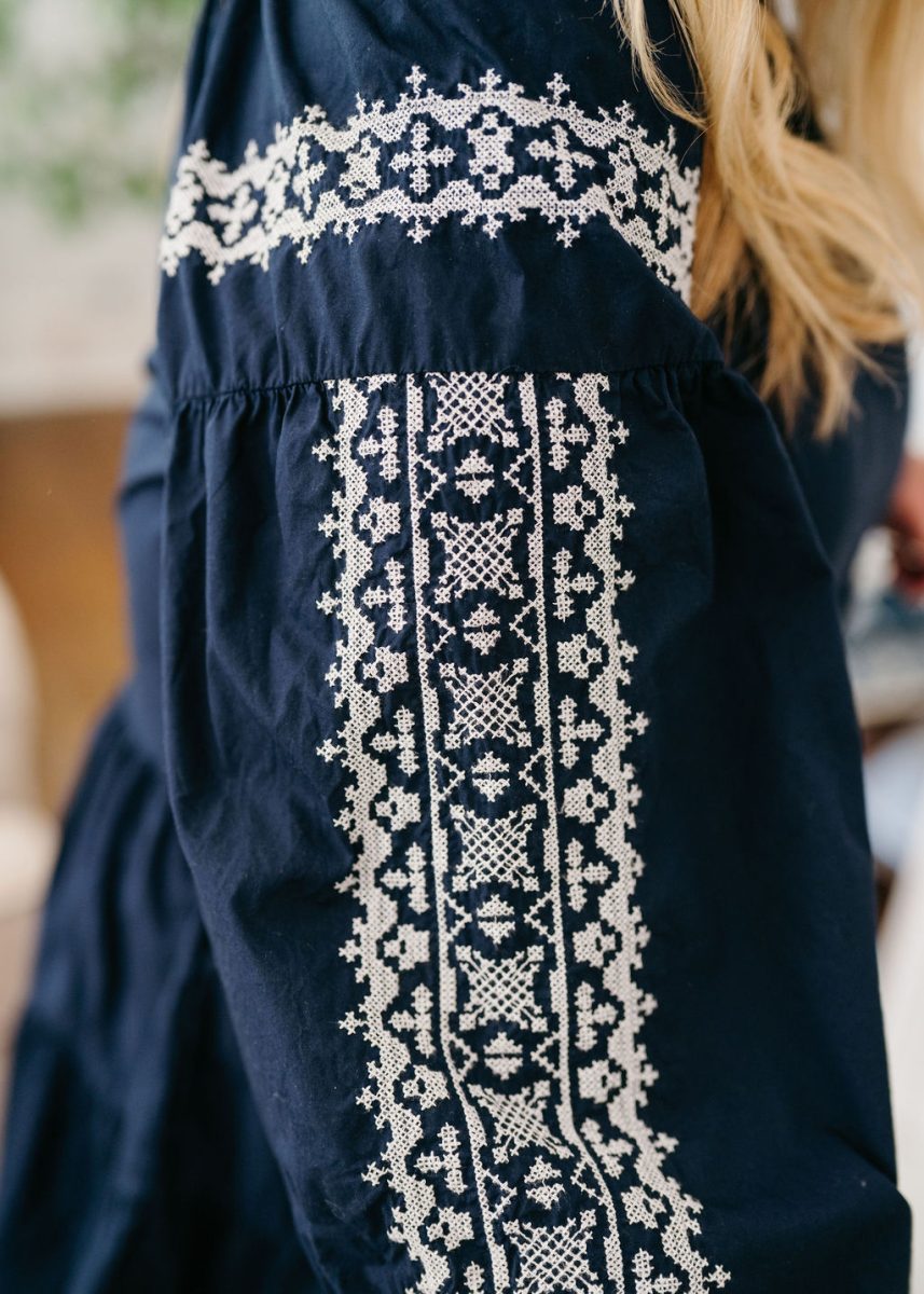 Close-up of a navy blue sleeve with intricate white embroidery patterns, worn by a person with long blonde hair. The background is softly blurred.