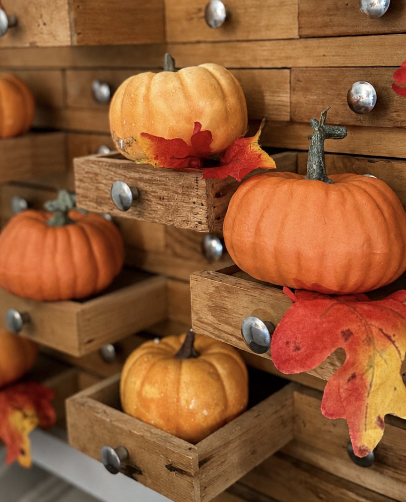 Small orange pumpkins and colorful autumn leaves placed on and inside open wooden drawers, creating a festive fall display.