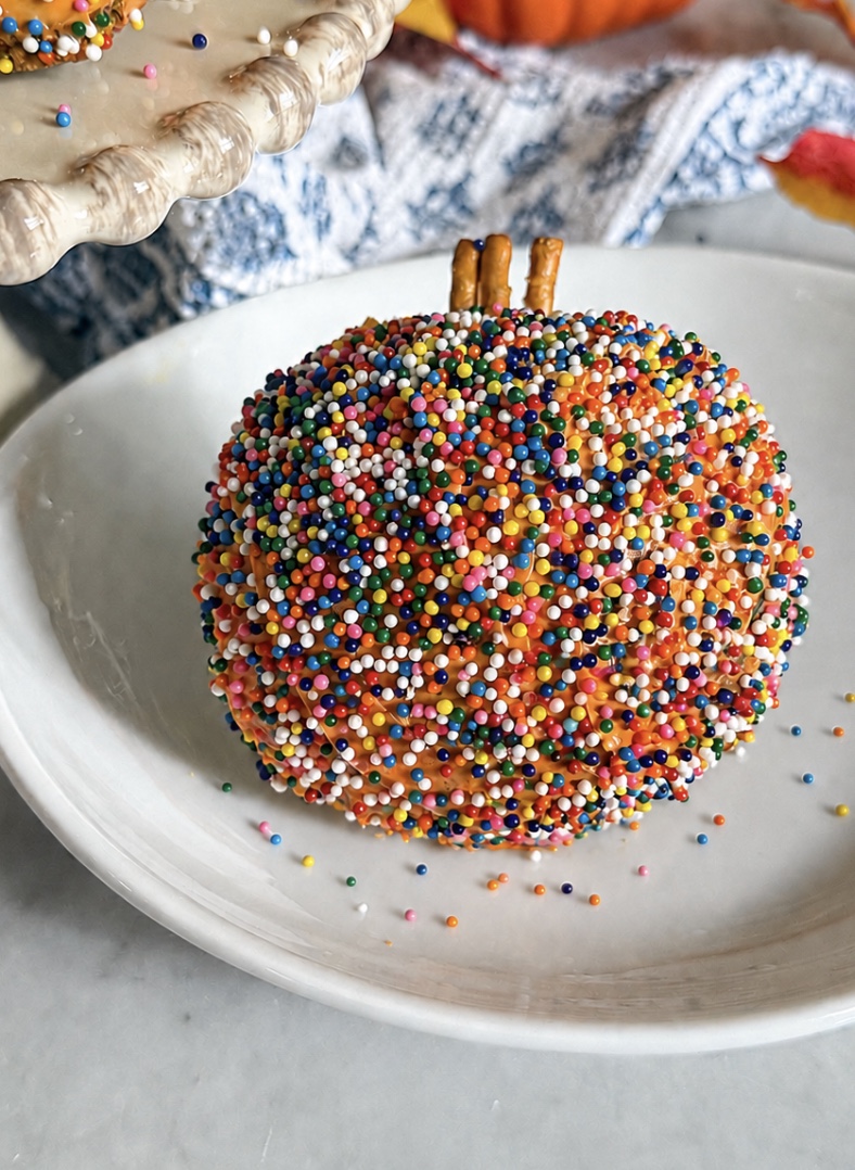 A round treat covered in colorful sprinkles sits on a white plate, topped with three pretzel sticks to resemble a pumpkin stem.