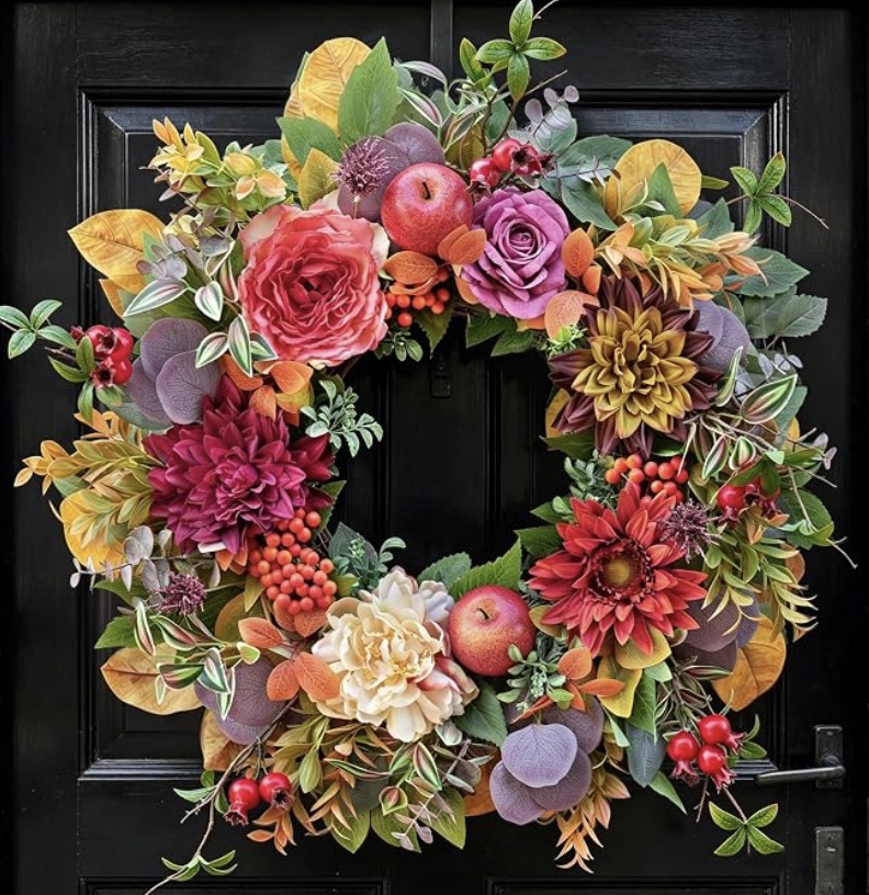 A decorative wreath with colorful flowers, leaves, berries, and apples hangs on a black door, featuring red, pink, and cream blooms with autumn foliage accents.