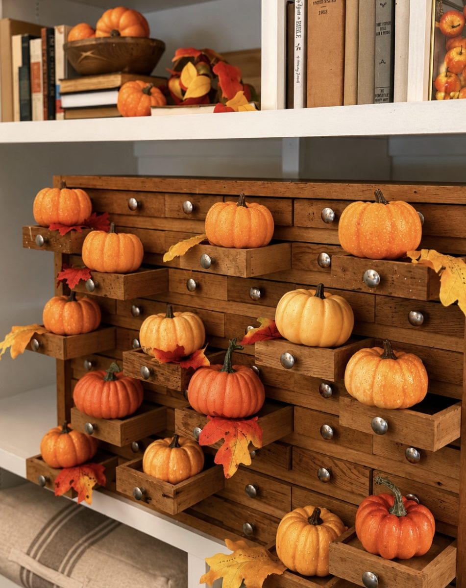 Small orange pumpkins and autumn leaves are displayed on the open drawers of a vintage wooden cabinet, with books and more pumpkins arranged on white shelves above.