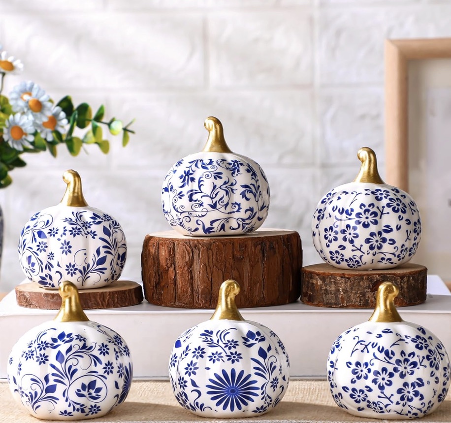 Six white ceramic decorative gourds with blue floral patterns and gold stems are arranged on wooden stands, with a plant and framed picture in the background.