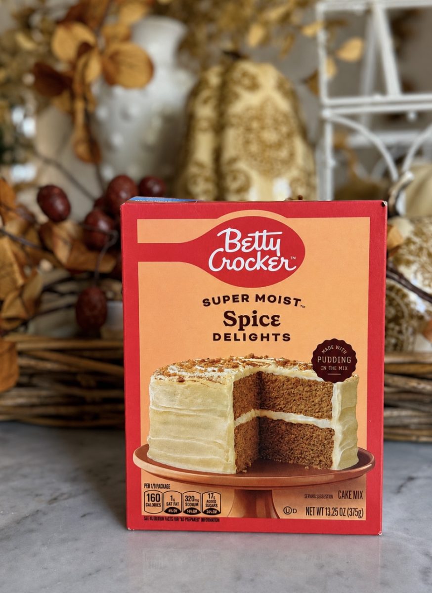 A box of Betty Crocker Super Moist Spice Delights cake mix sits on a counter, with dried fall foliage decor in the background. The box features an image of a frosted layer cake.