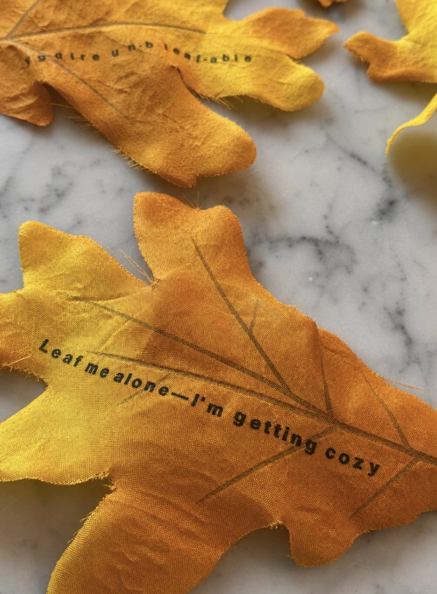 Yellow fabric leaves on a marble surface. One leaf has the text “Leaf me alone—I’m getting cozy” printed on it in black letters.
