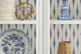 A white shelf displays blue and white ceramics, a wicker lamp, books, and gold decorative orbs, against a blue and white patterned wall.