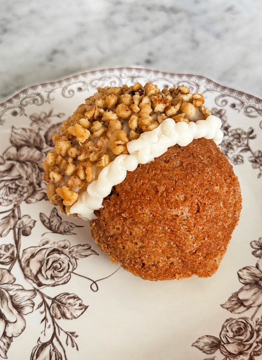 A cupcake decorated to look like an acorn, topped with chopped nuts and a band of white frosting, sits on a floral-patterned plate.
