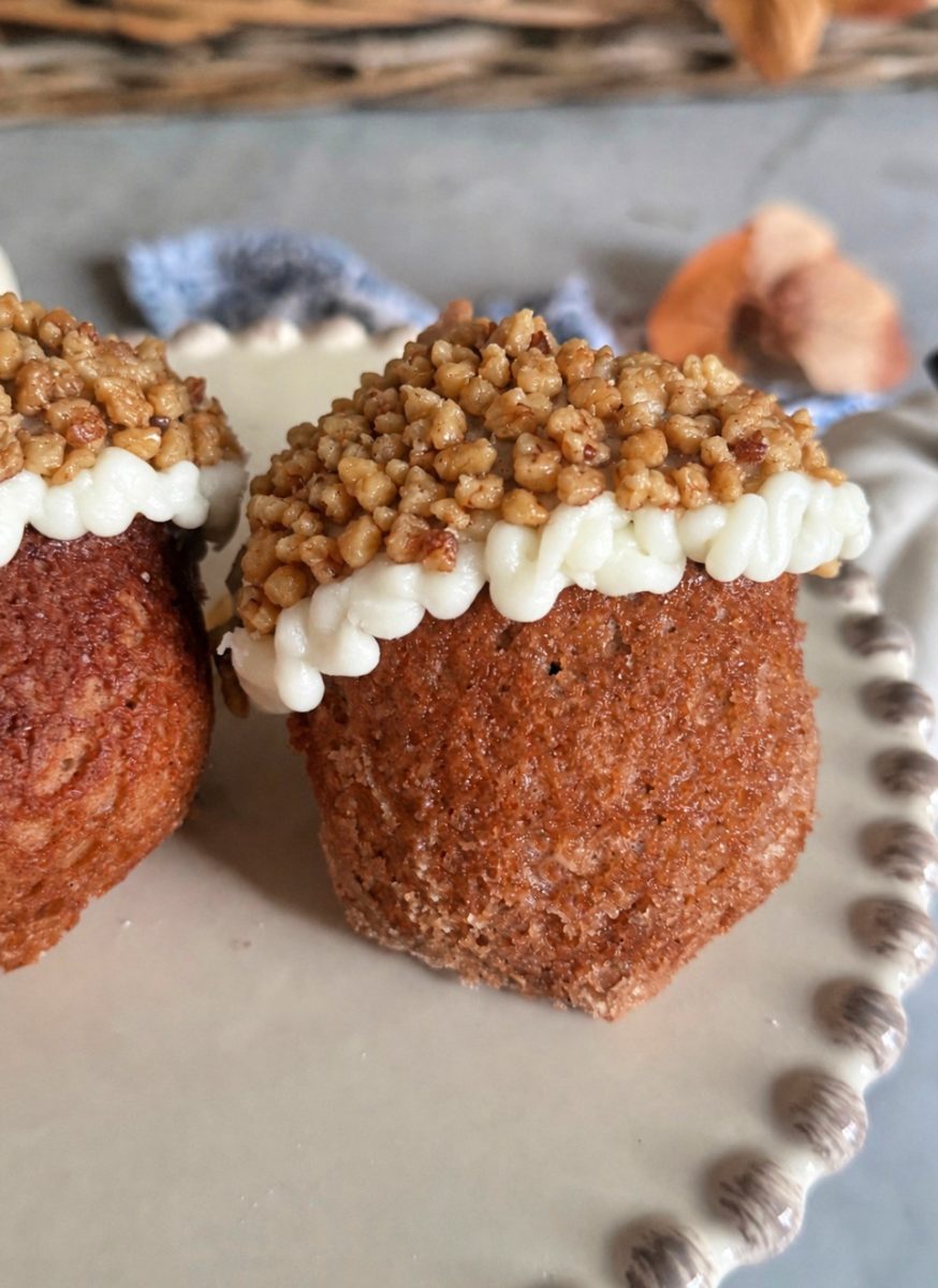 A close-up of a muffin topped with white icing and a layer of chopped nuts sits on a cream-colored plate with a beaded edge. Another muffin is partially visible beside it.
