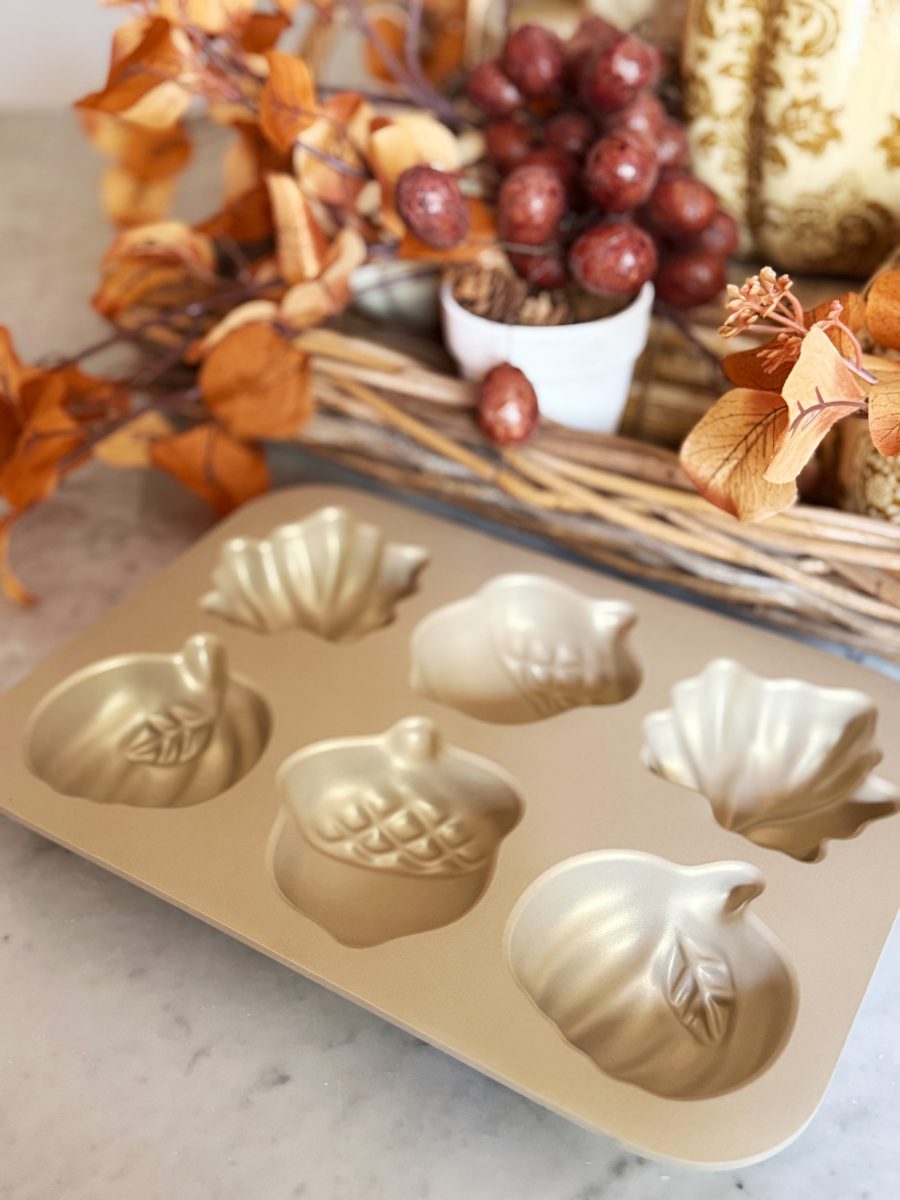 A gold baking pan with six autumn-themed molds sits on a marble counter, surrounded by fall decor including orange leaves, faux berries, and a wicker basket centerpiece.