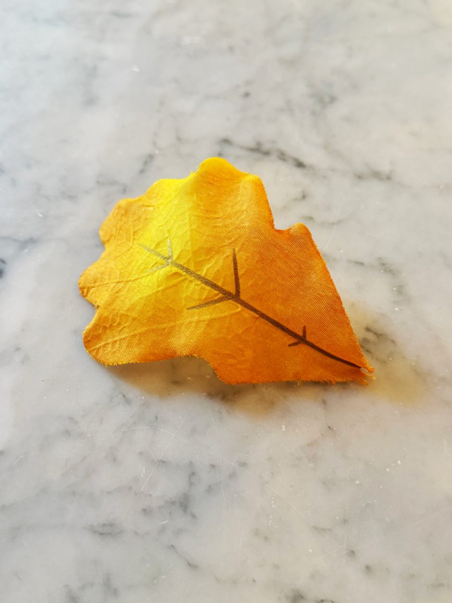 A single artificial yellow-orange leaf with brown veins lies on a light gray marble surface.