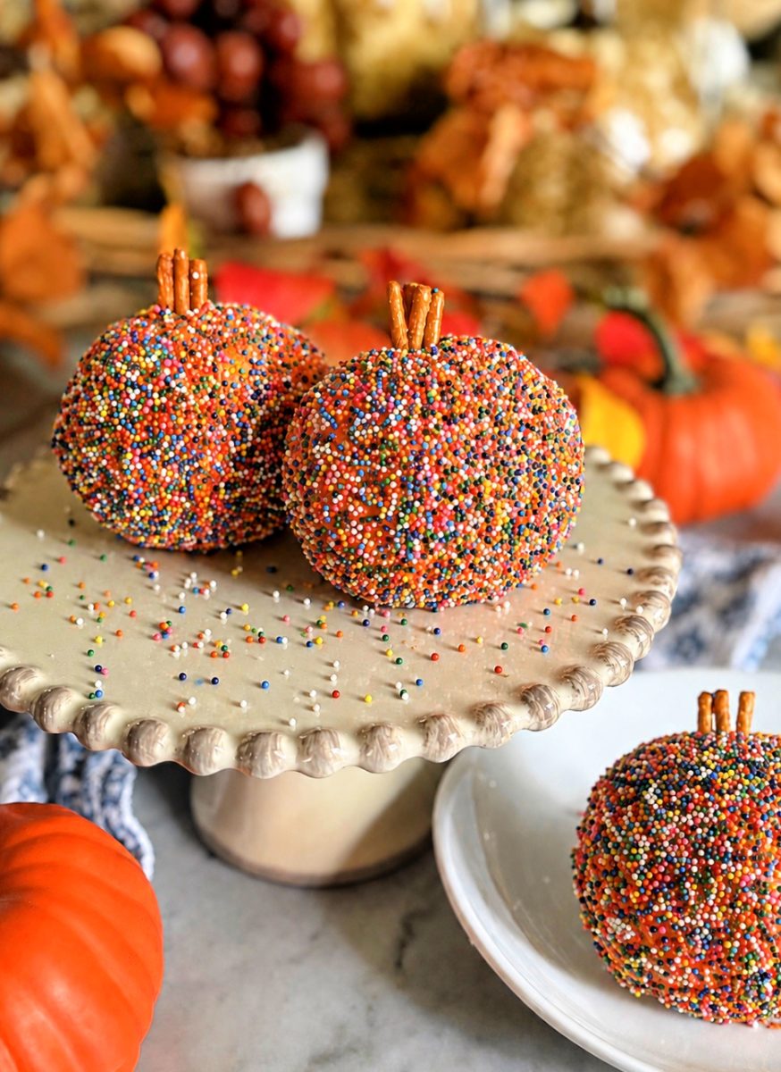 Three apple treats coated in rainbow sprinkles with pretzel stick stems sit on a stand and plate. Autumn decorations and pumpkins are blurred in the background.