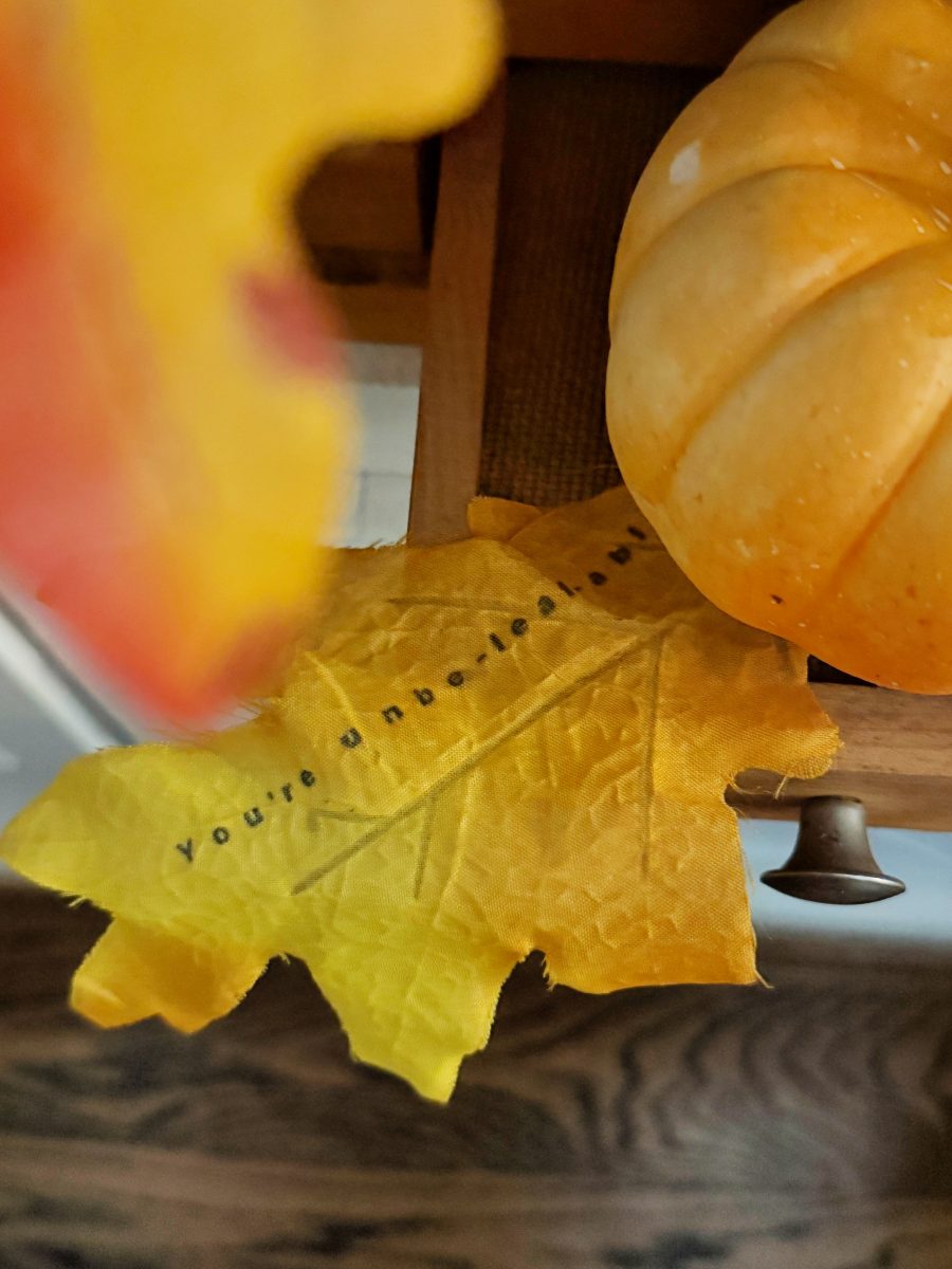 A yellow leaf with the text “You’re unbe-leaf-able” sits next to a small orange pumpkin on a wooden surface.