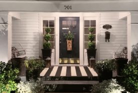 The front porch of a white house with a dark front door, black and white striped porch rug, and two chairs under a house number sign reading 701.