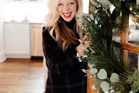 A smiling woman with blonde hair stands in a doorway decorated with a festive pine wreath.